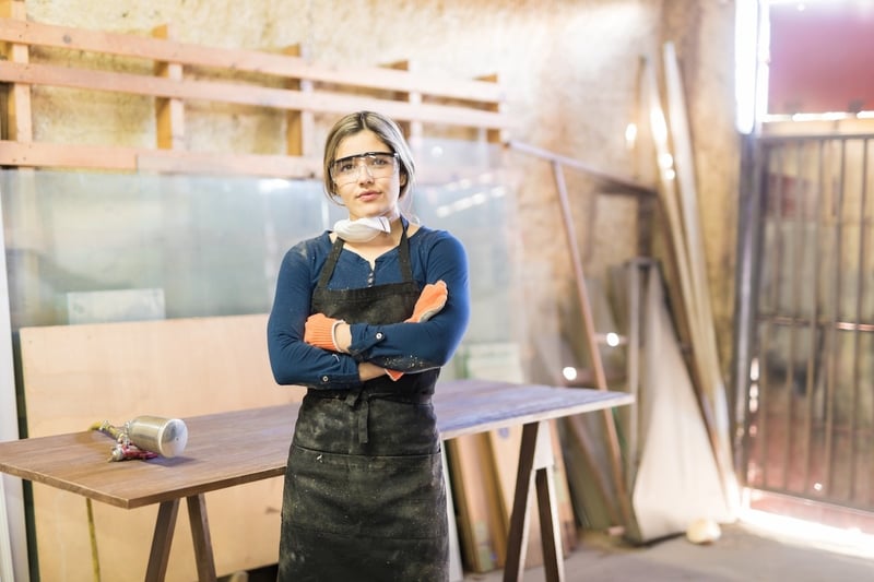 woodworker in workshop equipped with ppe facing camera with arms crossed next to unfinished wood and industrial sprayer